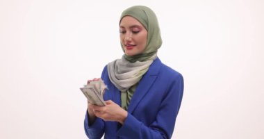 Mutlu Müslüman iş kadını beyaz arka planda nakit para tutuyor. Türbanlı kadın girişimci gelir miktarının yavaş çekimde memnuniyetini ifade ediyor
