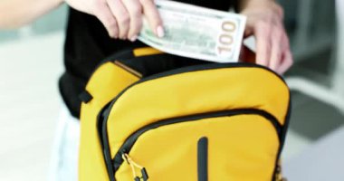 Parlak sarı bir sırt çantasına para koyan bir kişi gözlemlenebilir. Bu da finansal işlemleri açıkça simgeler.