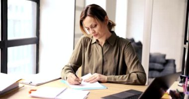 Bir kadın modern ofis masasına titiz bir şekilde detaylı notlar yazarak odaklanmış, doğal ışıkla kaplanmış.