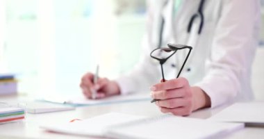 Çalışkan bir doktor tıbbi bir ofiste not alıyor ve gözlük tutuyor. Profesyonellik ve kendini adamışlık gösteriyor.