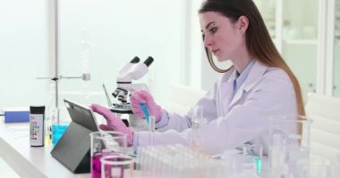 Kadın bilim adamı elinde mavi sıvı reaktörü içeren test tüpü tutuyor. Yoğunlaştırılmış araştırmacı yavaş çekim deneyinin sonuçlarını kaydetmek için tablet kullanır