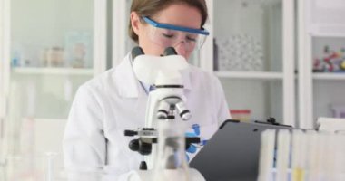 Kadın bilim adamı mikroskop kullanarak numunenin mikroskobik detaylarını inceliyor. Araştırmacı, analizlerin sonuçlarını kağıt defterde kaydeder.