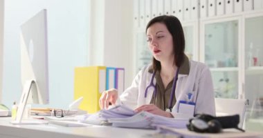 Yorgun kadın doktor klinikteki işyerinde kafaya dokunan kağıtları tutuyor. Üniformalı çok çalışan kadın doktor, tıbbi geçmişinde yorgunluk yaşıyor.