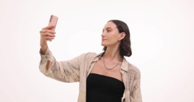 Genç bir kadın beyaz arka planda akıllı telefondan selfie çekerken poz veriyor. Hoş gülüşlü çekici kadın kişisel blog için içerik oluşturuyor