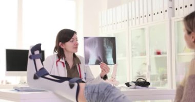 Kadın doktor, ayak yaralanmasının röntgen görüntüsünü muayenehanede tuttuğunu söylüyor. Bayan doktor ortopedik tedaviyi bacak korsesi takan kadın hastaya açıkladı.