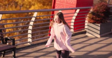 Uzun kollu kapüşonlu neşeli küçük kız yavaşça bankta oturan gülümseyen annesinin yanından geçiyor. Aktif çocuk, ebeveyniyle köprüde oynuyor