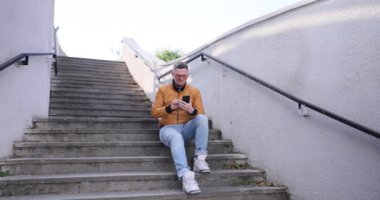 Modern bir şehir ortamında, şık bir genç adam merdivenlerde oturuyor, telefonunun keyfini çıkarıyor.