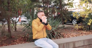 Bir insan açık havada oturmuş, canlı, renkli sonbahar yaprakları ve yemyeşil çimlerin ortasında hapşırıyor.