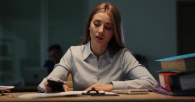 Yorgun kadın ofis çalışanı esneyerek akıllı telefonu kontrol ediyor ve iş yerinde el ele tutuşuyor. Bayan akşamları aşırı iş yükünden muzdarip.