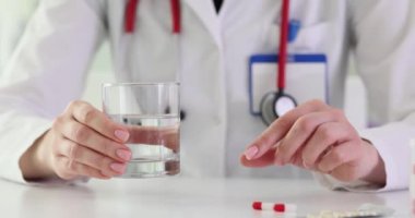 Kadın doktor elinde bir bardak su ve küçük ilaç kapsülü tutuyor. Doktorlar, ilaçların sorumluca alınmasının önemini vurguluyor. Yavaş çekim