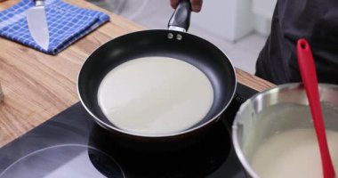 Mutfak işçisi mutfakta tavaya ince krep hamuru serpiyor. Adam ısıtılmış indüksiyon yüzeyinde yavaş çekimde krep pişiriyor.
