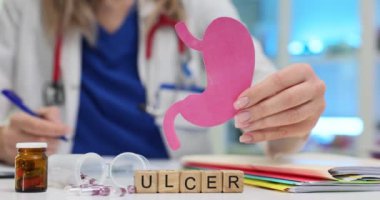 Küp üzerinde ülser ve masada kağıt mide gösteren bir doktor. Kadın tıp çalışanı, mide bağırsağında açık yaranın nedenlerini açıklıyor.