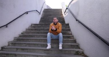 Adam sokak merdivenlerinde oturur, başını ezici duygularla tutar. Deri ceketli adam kol saatine bakıyor. Alnına elini koyuyor. Endişelerini dile getiriyor.