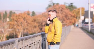 Palto giyen bir adam telefonda üstgeçit köprüsünde sonbahar ağaçlarına karşı konuşuyor. Adam hareketli konuşma sırasında heyecanını dışa vuruyor.
