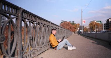 Adam kaldırımda dizüstü bilgisayarla üstgeçit köprüsü üzerinde çalışıyor. Güneş gözlüklü neşeli adam, sonbahar günü bilgisayarda yazan müşterilere cevap veriyor.