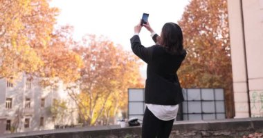 Kadın, sarı ağaçların arasında yaya bölgesinde dururken akıllı telefondan selfie çekiyor. Esmer kadın cep telefonuyla sosyal medya için içerik yaratırken anı yakalıyor