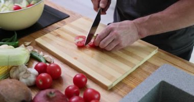 Adam kiraz domateslerini tahta kesme tahtasında bıçakla dilimliyor. Erkek aşçı sebze hazırlıyor. Ev mutfağında vejetaryen yemeği pişiriyor. Yavaş çekimde.