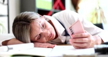 Masasındaki bir kadın, elinde telefon tutarken başını dinlendiriyor. İş yerinde stres ve yorgunluk belirtileri gösteriyor.