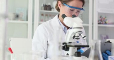 Laboratuvar çalışanı örnek özelliklerini mikroskop altında inceliyor. Kadın laboratuvar asistanı gözlemleri not defterine kaydeder. Yavaş çekim
