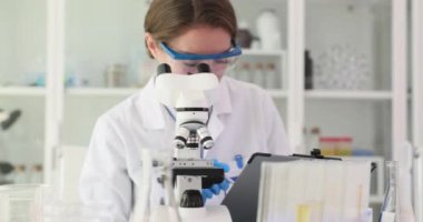 Gözlüklü bilim adamı örnekteki küçük değişiklikleri gözlemlemek için mikroskop kullanır. Konsantre kadın araştırmacı defterine sonuçlar yazar. Yavaş çekim