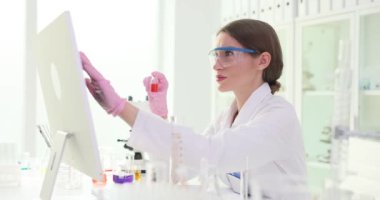 Kadın kimyagerin elinde kırmızı sıvıyla dolu bir kap var. Laboratuvar teknisyeni ayıraç hakkında daha fazla bilgi toplamak için bilgisayar kullanıyor. Yavaş çekim