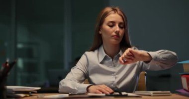 Yorgun kadın şirket çalışanı masasında esneyen bileğe bakıyor. Genç bayan şirket yöneticisi ofiste fazla mesai yaptığı için yorgunluk yaşıyor.