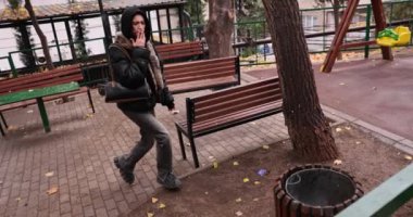 Young woman smokes cigarette near playground throwing litter into trash can. Brunette female puts out cigarette butt on bin side in city park in autumn