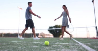 Bir çift gün batımında iyi aydınlatılmış bir kortta birlikte tenis oynuyor, atletizm ve keyif gösteriyorlar.