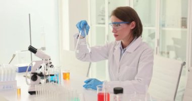 Kadın bilim adamı Erlenmeyer şişesindeki şeffaf sıvıyı modern laboratuvar ortamında inceliyor. Asistanı masadaki cam kabın içinde suyu sallıyor.