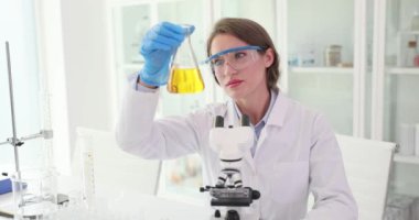 Kadın bilim adamı masasındaki Erlenmeyer matarasındaki sarı sıvıya bakıyor. Odaklanmış kimyager, tıp laboratuarında çalışan numune içeriğini inceliyor.