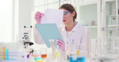 Güvenlik gözlüklü kadın bilim adamı laboratuvarda yavaş çekimde notlar okuyor. Test tüpleri mikroskobu ve ekipmanlar tıbbi laboratuarda düzenlendi.