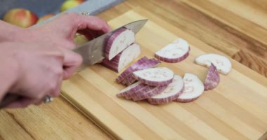 Kadın şef, tahta kesme tahtasına graffiti patlıcan dilimliyor. Kadın, mutfağın aydınlatması için yavaş çekimde taze malzeme hazırlıyor.