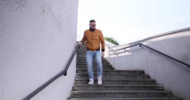 Adam yer altındaki beton merdivenlerden aşağı inip kol saatini kontrol ediyor. Güneş gözlüklü genç adam çevreye bakıyor ve ağır çekimde gezinmeye çalışıyor.