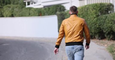 Kısa saçlı genç adam kaldırımların yanındaki sık çalılarla boş cadde boyunca yürüyor. Şık giyimli bir adam yerleşim bölgesinde ağır çekimde geziniyor.