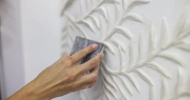 Kadın sanatçı, beyaz duvarda dikkatlice kumlu alçı yaprak desenli. Tasarımcı yavaş çekimde sanatın pürüzsüz yüzeyi için aşındırıcı sünger kullanır