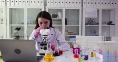 Bayan araştırmacı laboratuvar örneklerini mikroskop altında inceler ve bilgisayarla sonuç verir. Kadın kimyager araştırma alanında renkli kimyasalların arasında çalışıyor.