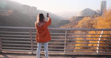 Kadın yaya köprüsünde dururken güzel sonbahar kentsel manzarasını akıllı telefonuyla yakalıyor. Turist günbatımında şehrin mevsimlik güzelliğinin fotoğrafını çekiyor.