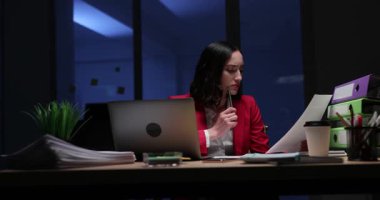 Düşünceli iş kadını akşamları iş yerinde dizüstü bilgisayarıyla oturan kağıt belgeleri okur. Ofiste çalışan odaklı kadın kontrolleri mali rapor