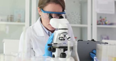 Kadın laboratuvar çalışanı, iş yerinde mikroskop kullanarak örnek alıyor. Gözlüklü ve üniformalı kadın bilim adamı hayati noktaları vurgulayan notlar alıyor.