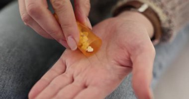 Kadın evdeki koltukta oturan şişeden avuç içine beyaz haplar döküyor. Kadın, hastalıkla başa çıkmak için doğru dozda ilaç hazırlıyor.