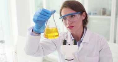 Kadın bilim adamı Erlenmeyer şişesindeki sarı sıvıyı mikroskop yakınında inceliyor. Kimyager modern laboratuvar ortamında zeytinyağı araştırması yapıyor.