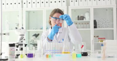 Kadın bilim adamı laboratuarda kırmızı sıvı içeren test tüpünü inceliyor. Kimyager, iş yerinde sıvı reaksiyonuyla şişeyi vurur ve sallar.