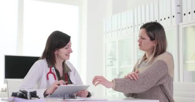 Kadın doktor, teşhis konsültasyonunda bilek rahatsızlığı çeken bir kadını dinliyor. Sıhhiye klinikteki semptomları panoya belgeledi