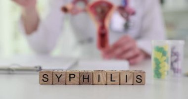 Jinekolog, hastalığın ilerleyişini göstermek için yapay uterus modeli kullanır. Tahta bloklar doktor masasında SyPHILIS kelimesi oluşturur. Yavaş çekim