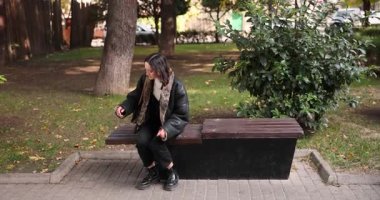 Genç bir kadın oturarak kol saatindeki zamanı kontrol ediyor. Deri ceketli bir kadın, şehir parkında oturan yeşil yavaş çekimde arkadaşını bekliyor.