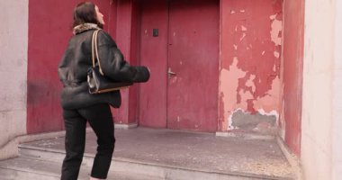 Deri ceketli genç bir kadın pejmürde giriş kapısına ağır çekimde yürür. Çantalı kadın dikkatli bir şekilde kapıyı çalıyor ve ev sahibinden yanıt bekliyor.