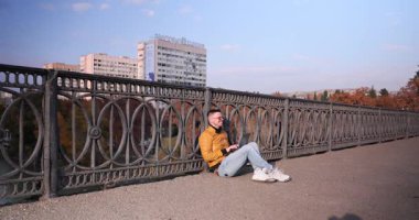 Ayakköprüsü zemininde oturan gülümseyen genç adam dizüstü bilgisayarda yazı yazıyor. Deri ceketli genç adam sıcak sonbahar gününün tadını çıkarıyor.