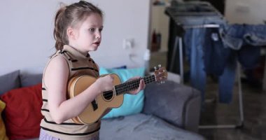 Ukulele çalan küçük kız oturma odasında tutkuyla şarkı söylüyor. Pozitif çocuk evde ağır çekimde telli çalgılarla melodi çalıyor