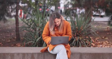 Endişeli kadın, şehir parkında parapet üzerinde oturan dizüstü bilgisayarla çalışıyor. Kadın ofis çalışanı Autumn City caddesinde dizüstü bilgisayar kullanıyor.