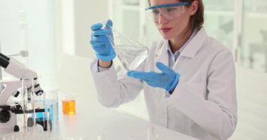 Kadın teknisyen, laboratuvar ortamında Erlenmeyer şişesinde temiz sıvı gözlemliyor. Kimyager araştırma sırasında su kalitesini kontrol ediyor.
